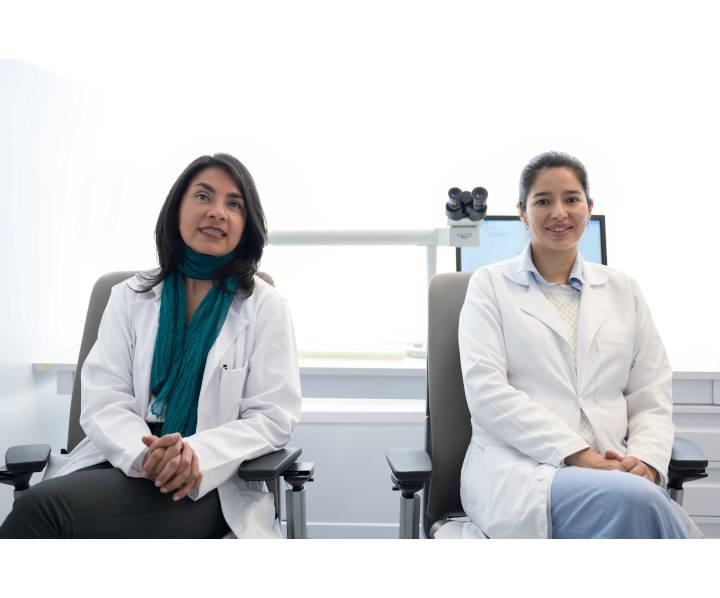 Dos doctoras se encuentran sentadas posando frente a la cámara sonriendo.