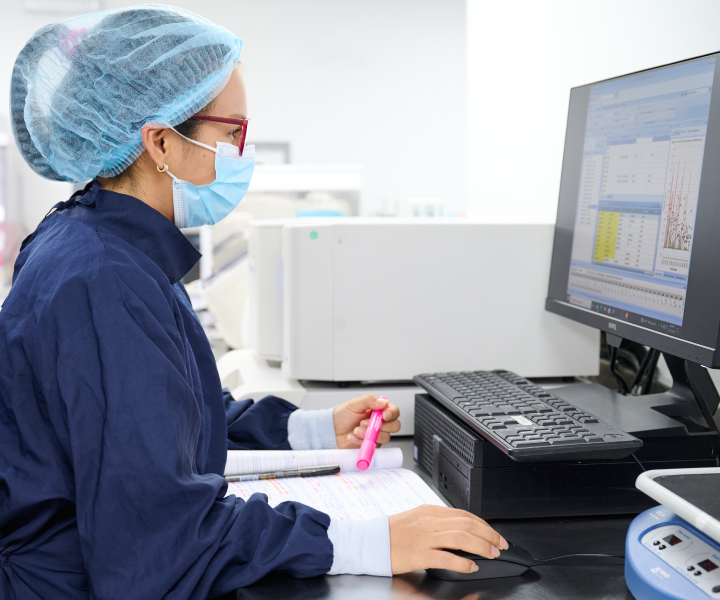 Una profesional de la salud con gorro y tapabocas se encuentra sentada frente a una computadora revisando resultados de laboratorio.