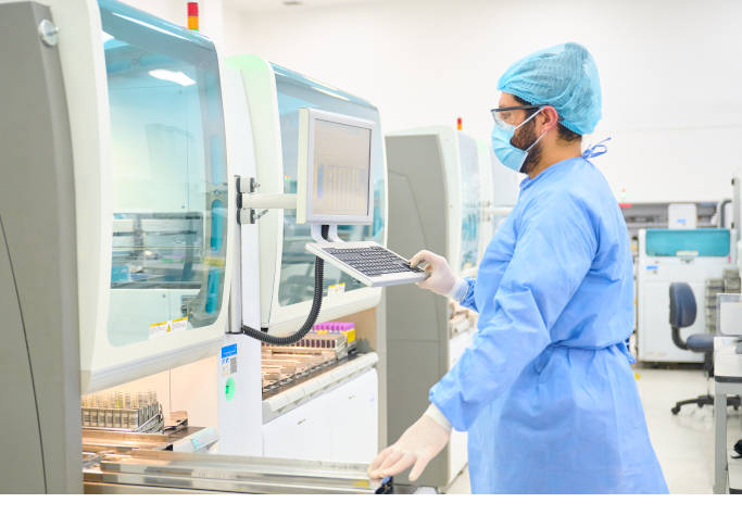 En un ambiente de laboratorio se ve a un profesional de la salud, de pie, vestido con equipo de protección médico, operando un computador que está anclado a una pared. 