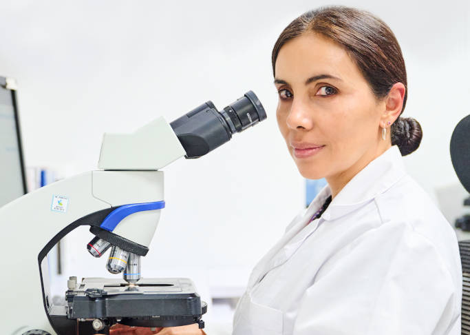 Se ve a una profesional de la salud sentada  mirando al frente a la cámara, frente a su cara hay un microscopio. 