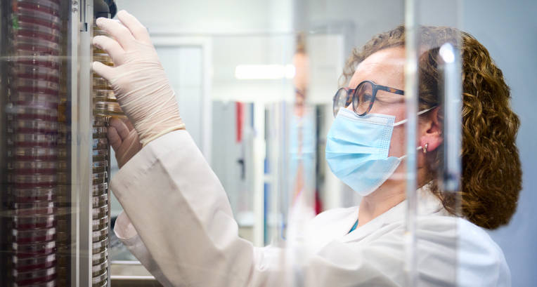 Se ve en primer plano a una mujer que se puede inferir que es profesional de la salud usa tapabocas y guantes y está mirando fijamente un objeto que por el encuadre se pierde del plano. 