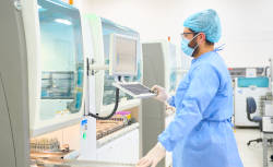 En un ambiente de laboratorio se ve a un profesional de la salud, de pie, vestido con equipo de protección médico, operando un computador que está ...