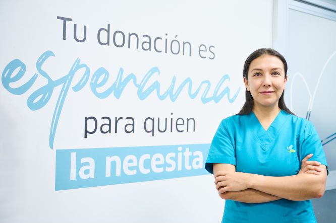 Una profesional de la salud se encuentra posando de pie con los brazos cruzados frente a un mensaje  en la pared invitando a donar sangre.