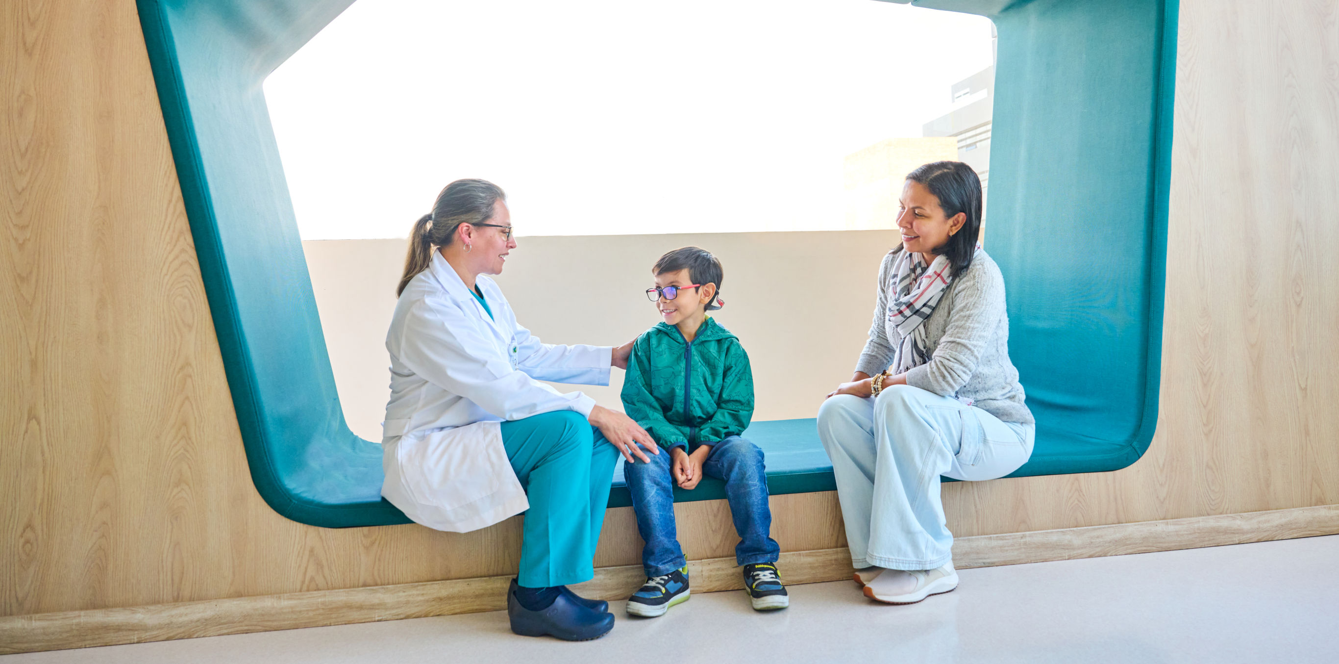  En un ambiente de consultorio hay una profesional de la salud, un niño y una mujer, los 3 están interactuando en una conversación que se puede inferir como positiva, porque están sonriendo. 