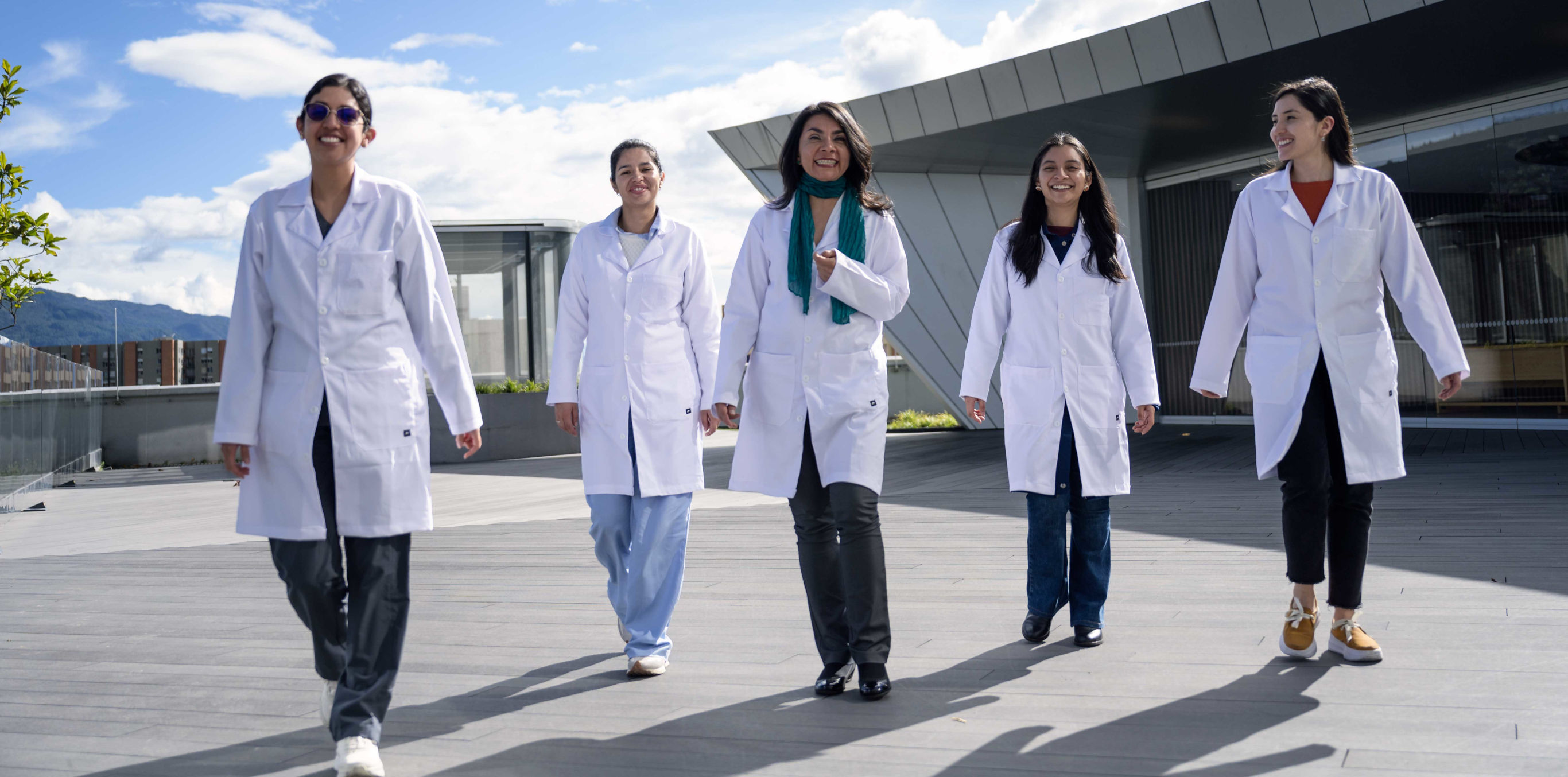 Se observan 5 profesionales de la salud de pie sonriendo y caminando hacia la cámara frente una sede de clínica.