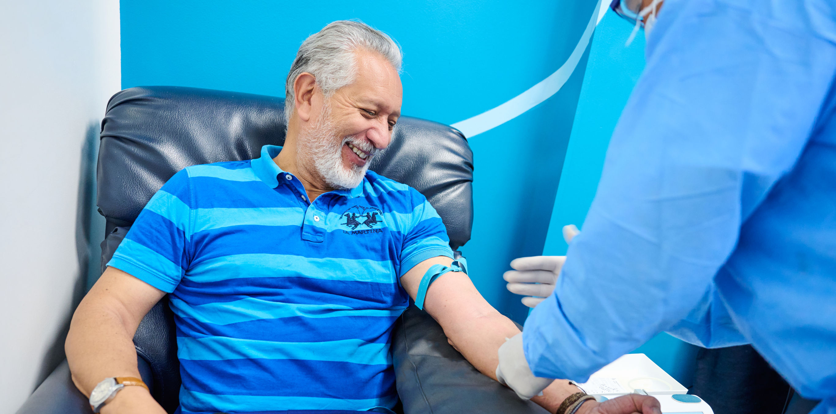 Un adulto mayor está sentado en la silla sonriendo mientras un profesional de la salud se encuentra tomando su brazo para tomar una muestra de laboratorio.