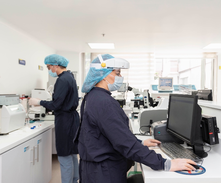se observa a dos doctoras, la primera se encuentra analizando muestras de laboratorio y la segunda sentada a la derecha consultando resultados en e...